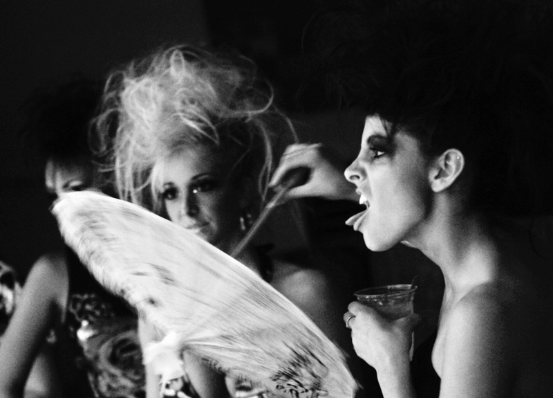 Black-and-white photo of models backstage at the Ed Hardy fashion show at Mantra, Boston, 2009: one fans herself with a feather fan while another sticks out her tongue, drink in hand