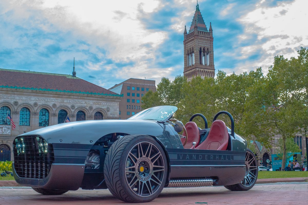 A grey Vanderhall three-wheeled roadster with red seats parked in Copley Square, Boston, with the Boston Public Library and Old South Church tower behind it, 2017